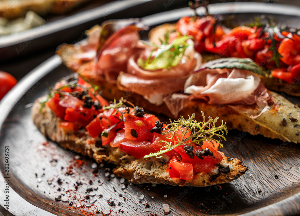 Variety of bruschetta with prosciutto, tomatoes, parmesan cheese, sauce pesto, grilled pepper on ciabatta bread on wooden plate over dark background. Sandwiches set of wine, close up