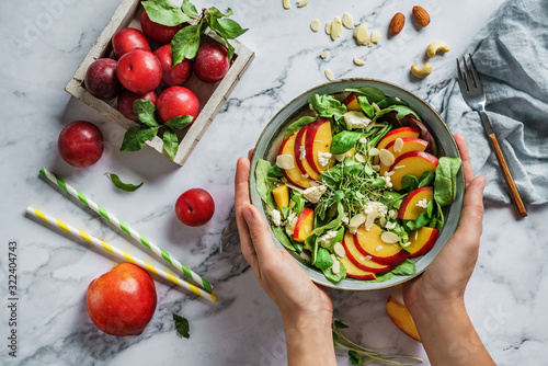 Hands holding fresh summer salad with peach, spinach, micro greens, plums, feta cheese and almonds on light marble background. Healthy food, clean eating, Buddha bowl salad, top view © Jukov studio