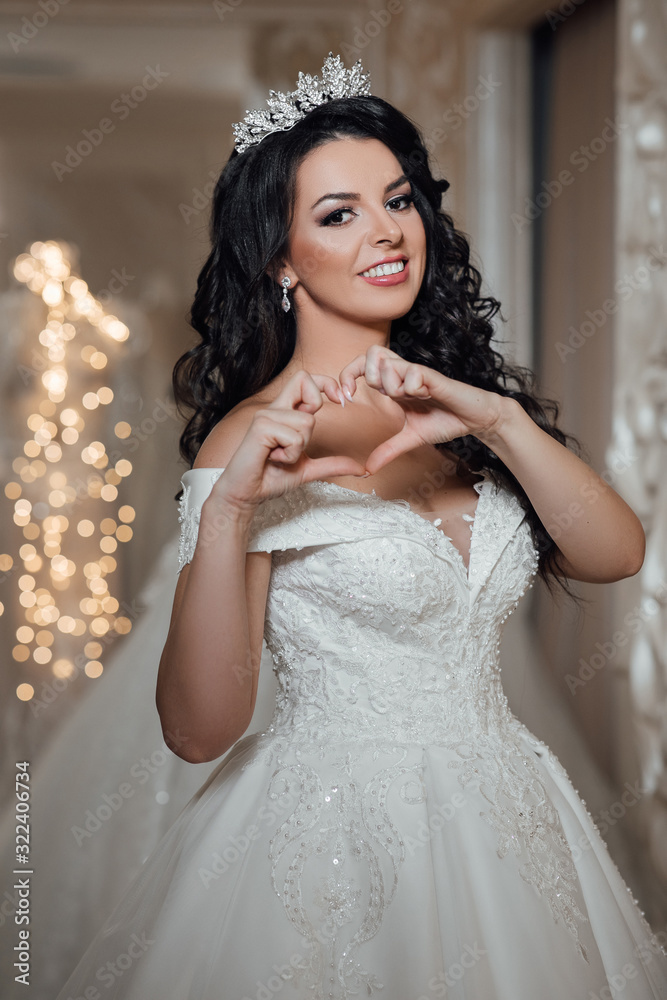 Wedding hairstyle and make up. The girl folded her heart. Smiling pretty bride chooses white gown at shop of wedding fashion. Young and beautiful bride posing in wedding dress. Wedding hairstyle and make up. The girl folded her heart. Smiling pretty bride chooses white gown at shop of wedding fashion. Young and beautiful bride posing in wedding dress.