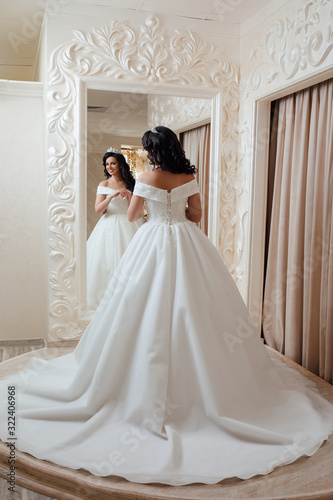 Beautiful young bride is looking into the mirror and smiling while choosing elegant wedding dresses in modern wedding salon. Wedding hairstyle and make up. 