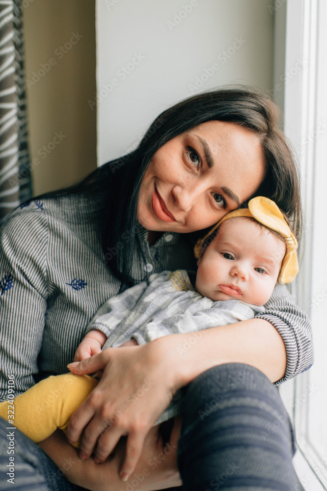 Portrait of beautiful woman with black hair sits on a windowsill with her pretty little child and smiles Portrait of beautiful woman with black hair sits on a windowsill with her pretty little child and smiles