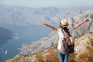 Traveler with backpack on mountain top. Happy woman with raised arms in amazing landscape. Girl tourist enjoying travel, adventure, freedom, vacation. Concept of solo female tourism, trip. Rear view.