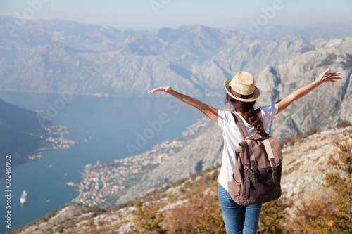 Traveler with backpack on mountain top. Happy woman with raised arms in amazing landscape. Girl tourist enjoying travel, adventure, freedom, vacation. Concept of solo female tourism, trip. Rear view.