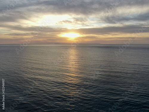 Aerial view of Pacific ocean colorful sunset, facing the sun with reflection on the ocean and deep clouded sky. Newport Beach, California