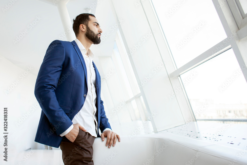 Successful handsome businessman in a blue suit at the workplace looks out the window, portrait of a manager at work Successful handsome businessman in a blue suit at the workplace looks out the window, portrait of a manager at work