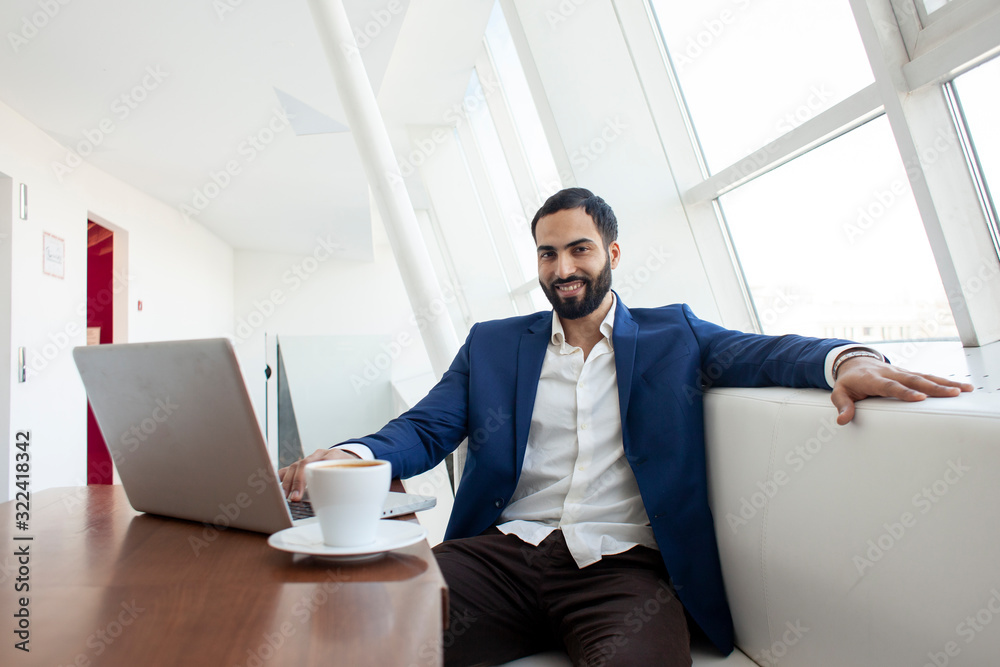 Fototapeta premium young successful businessman use a laptop and drink coffee in the morning at work, a freelancer is sitting with a computer in a white cafe
