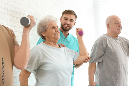 Care worker helping elderly woman to do exercise with dumbbell in hospital gym