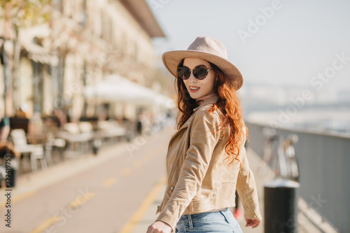 Portrait of laughing ginger woman looking over shoulder while walking down the street. Photo of joyful red-haired lady in sunglasses chilling at embankment.