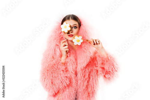 girl with candy on white in pink fur coat. Prettywoman with makeup attractively posing with lollipop on stick in studio.candy like chamomile, flower in hand.Joyful laughing lady at the party photo