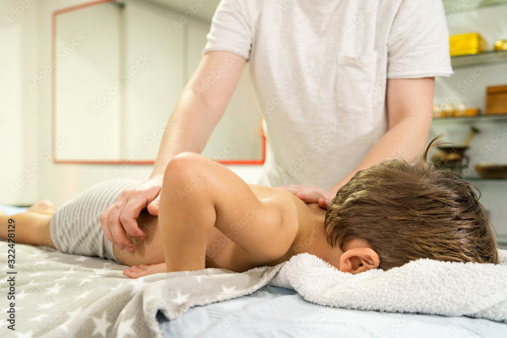 Small caucasian boy laying on the bed naked having back massage by professional female physiotherapist masseuse in a rehabilitation center salon or home office of physical therapy side view Small caucasian boy laying on the bed naked having back massage by professional female physiotherapist masseuse in a rehabilitation center salon or home office of physical therapy side view