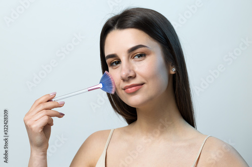 Close-up of a young girl in a light top on a white background making a facial make-up. A pretty woman holds a cosmetic brush near her face and smiles.