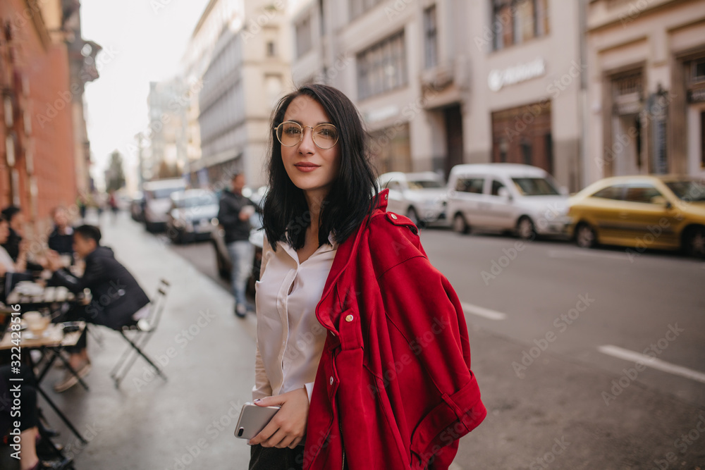 Pleased caucasian girl in glasses standing on the street with phone in hand. Smiling brunette young lady in red jacket posing with pleasure in city.