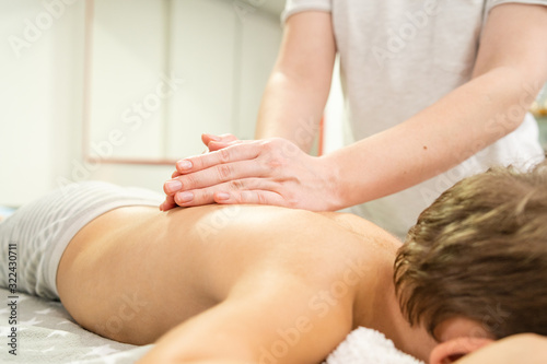 Small caucasian boy laying on the bed naked having back massage by professional female physiotherapist masseuse in a rehabilitation center salon or home office of physical therapy close up on hands