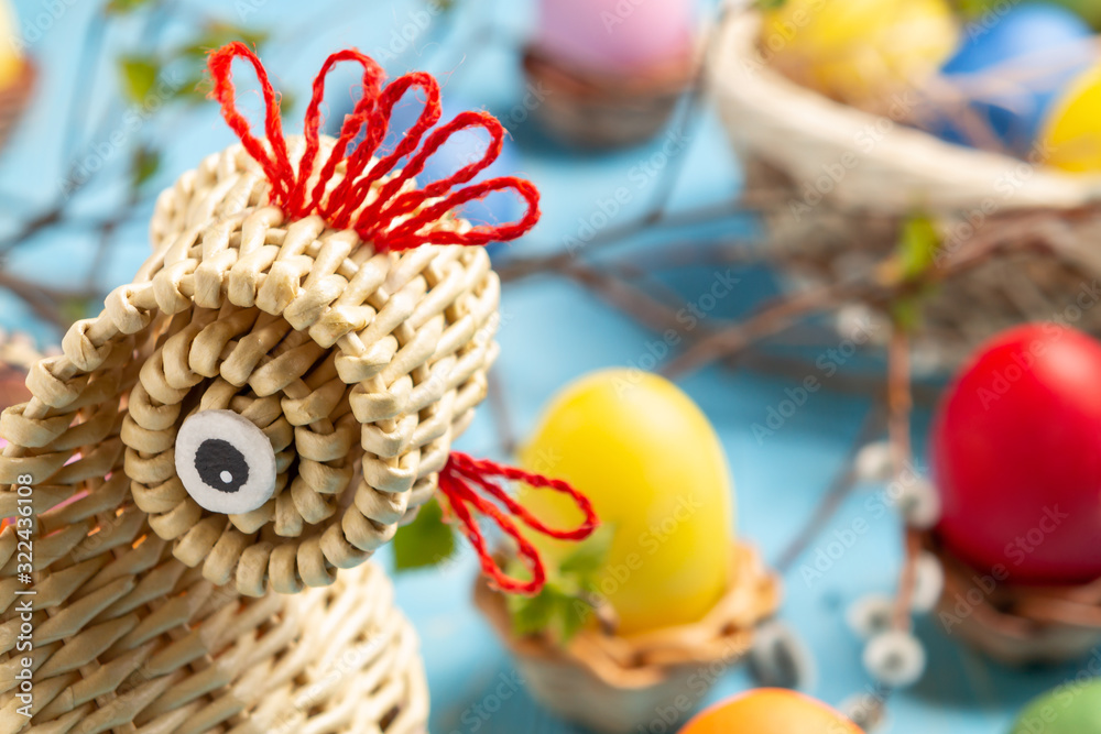 Easter composition - a wicker basket in the form of a chicken with colored eggs on a blue wooden table with willow twigs Easter composition - a wicker basket in the form of a chicken with colored eggs on a blue wooden table with willow twigs