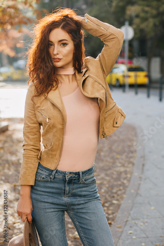 Slim ginger woman plays with her hair while posing in autumn day. Outdoor portrait of good-looking red-haired girl in blue jeans standing in park. © Look!