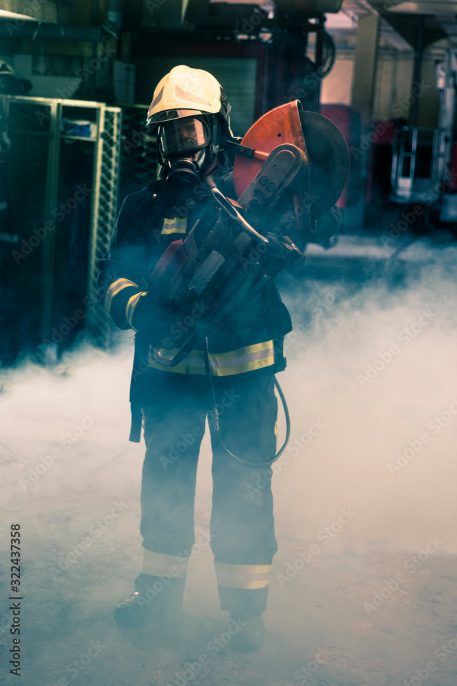 Portrait of a firefighter wearing full protective equipment posing with a chainsaw on his shoulder. Dark background with smoke and blue light. Portrait of a firefighter wearing full protective equipment posing with a chainsaw on his shoulder. Dark background with smoke and blue light.
