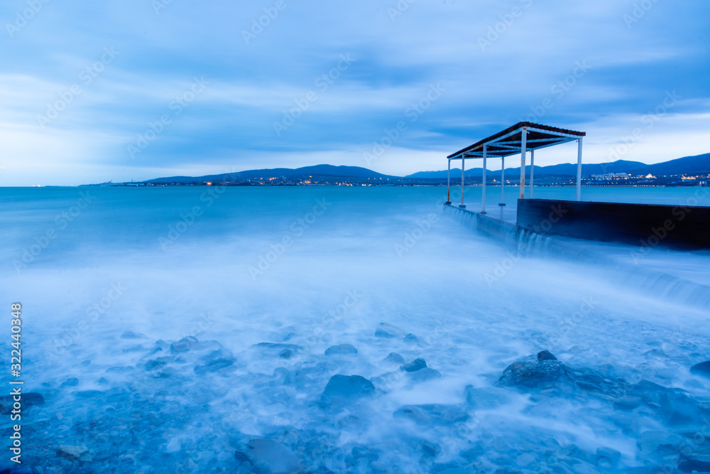 Storm waves at high speed run over the stones of Gelendzhik beach. Waves due to exposure are a blue fog. The resort of Gelendzhik, the evening twilight. 