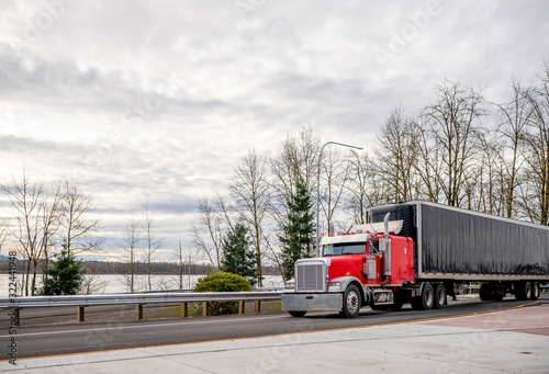 Big rig red classic bonnet semi truck transporting cargo in covered rubberized black semi trailer driving on the road intersection along the river