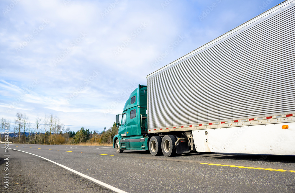Big rig semi truck with corrugated refrigerated semi trailer with aerodynamic skirt moving on the road with trees on the side Big rig semi truck with corrugated refrigerated semi trailer with aerodynamic skirt moving on the road with trees on the side