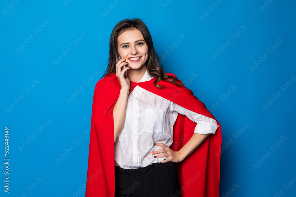 Fototapeta premium Business, power and people concept. Young smiling businesswoman in red superhero talk on the phone on blue background
