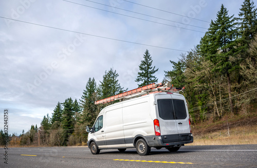 Small compact commercial cargo mini van with ladders on the roof running on the road with trees on the hill.