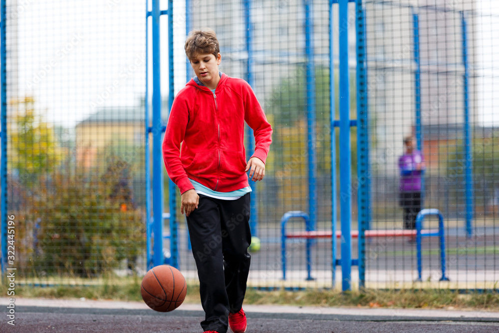 Cute smiling boy in red shirt plays basketball on city playground. Active teen enjoying outdoor game with orange ball. Hobby, active lifestyle, sport for kids. Cute smiling boy in red shirt plays basketball on city playground. Active teen enjoying outdoor game with orange ball. Hobby, active lifestyle, sport for kids.