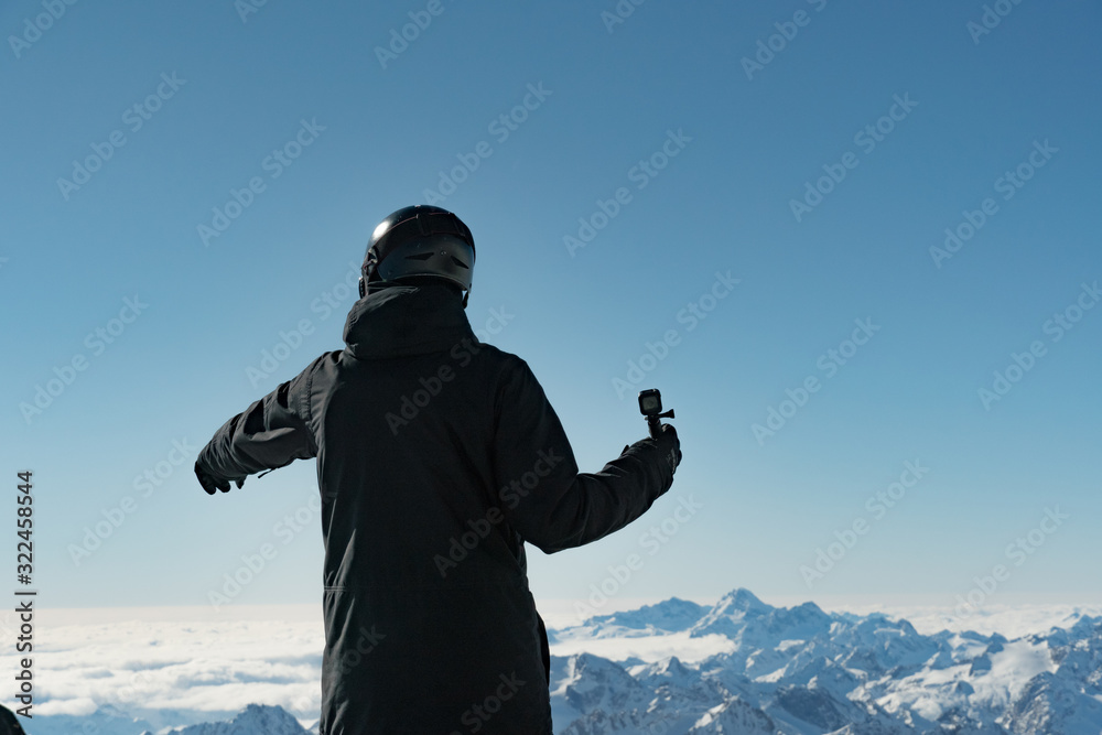 Male snowboarder in protective helmet standing on top of the mountain filming himself on action camera. Caucasus mountains in the background. Extreme winter sports. Male snowboarder in protective helmet standing on top of the mountain filming himself on action camera. Caucasus mountains in the background. Extreme winter sports.