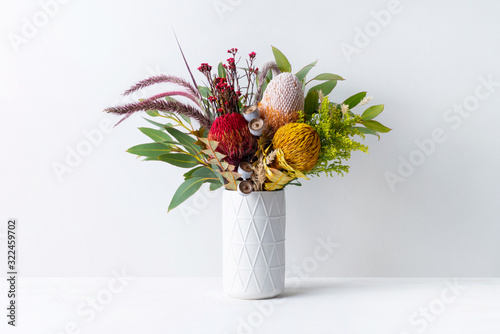 Fototapeta Naklejka Na Ścianę i Meble -  Beautiful floral arrangement of mostly Australian native banksias, eucalyptus leaves and gum nuts, fountain grass and dried kangaroo grass, in a white vase on a white table with a white background.
