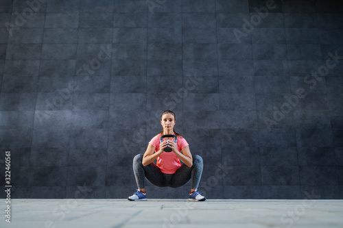 Attractive Caucasian brunette in sportswear doing squats endurance with kettle bell. In background is dark wall