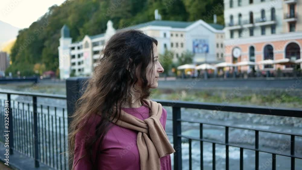 Beautiful young woman walking along river embankment of resort town in mountains Beautiful young woman walking along river embankment of resort town in mountains