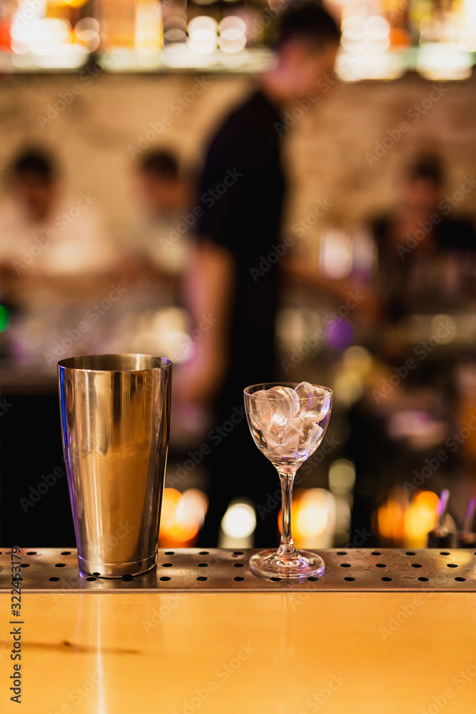 A nick and nora glass filled with ice and a shaker on a bar counter, people in bokeh, bar atmosphere