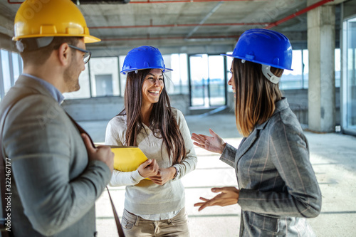 Group of young highly motivates attractive caucasian architects with helmets on heads standing in building in construction process and having casual talk on pause.