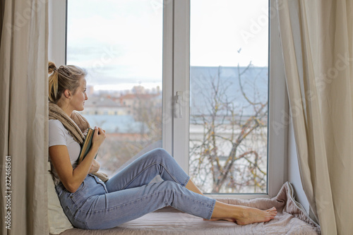 Beautiful young girl sitting on a windowsill with a book and dreamily looking out the window