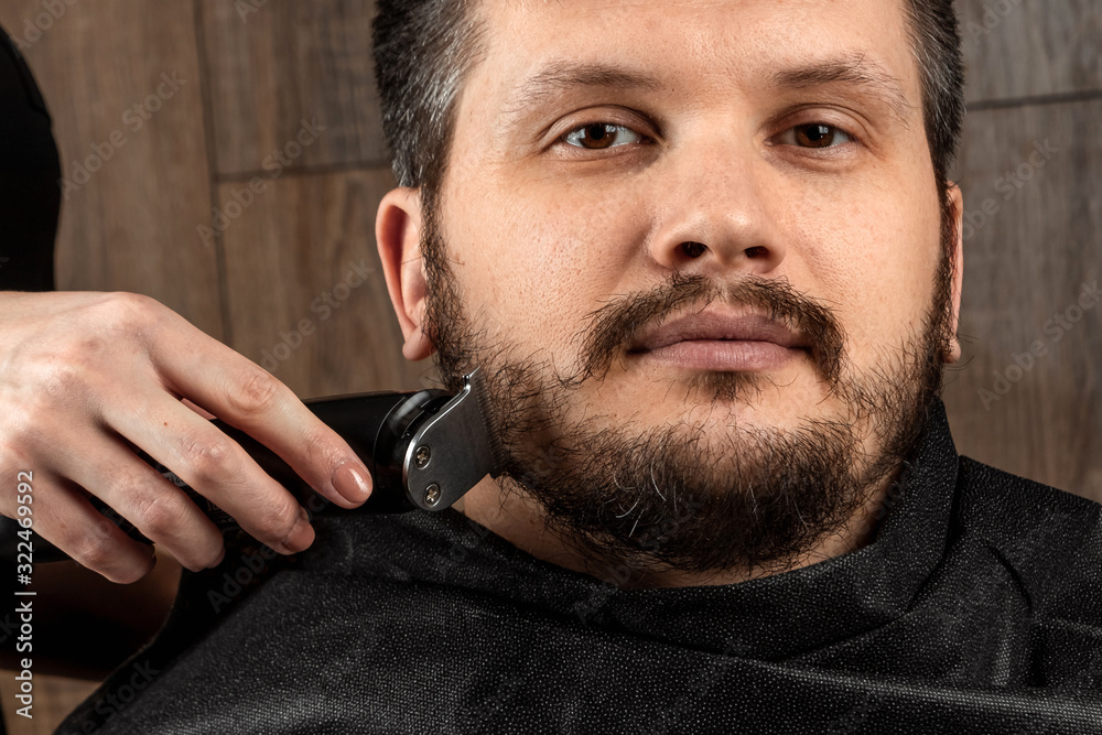 Obraz premium The master makes a beard haircut to the client with an electric machine, trimmer, close-up. The process of shaving a beard at the hairdresser. Body care, lifestyle, metrosexual.