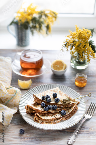 A plate with pancakes with blueberry berries on a wooden table. In the background is a cup of tea and a bouquet of spring flowers by the window.