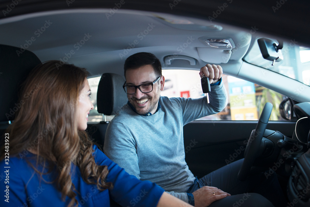 Young couple buying their first new vehicle at car dealership. Man holding car keys and woman happy and smiling while they are sitting in comfortable car interior.