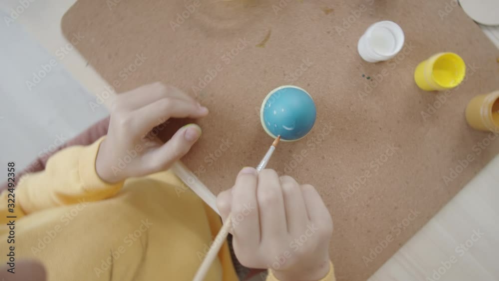 Top view shot of little girl applying dots with paint brush on blue dyed egg while decorating food for Easter
