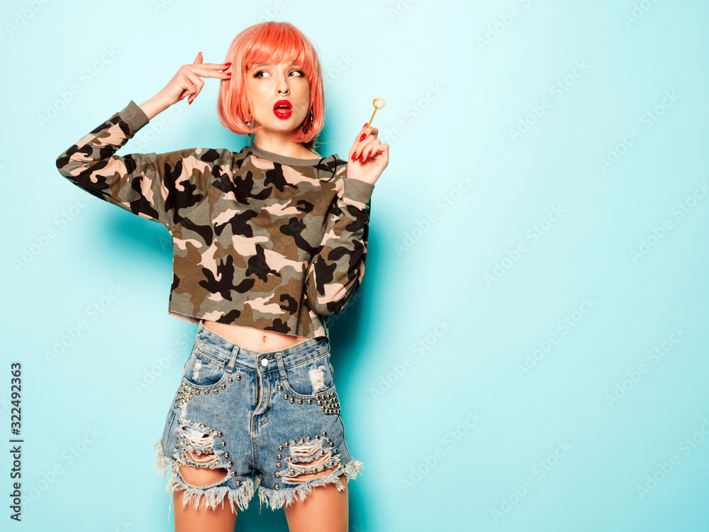 Portrait of young beautiful hipster bad girl in trendy jeans shorts and earring in her nose.Sexy carefree smiling woman in pink wig posing.Positive model licking round sugar candy.Makes gun sign
