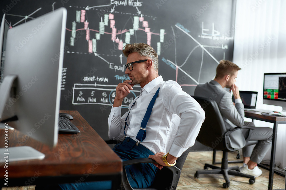 A Taste For Trading. Middle-aged trader sitting by desk in front of computer monitor while working in the office. His colleague and blackboard full of charts and data analyses in background. A Taste For Trading. Middle-aged trader sitting by desk in front of computer monitor while working in the office. His colleague and blackboard full of charts and data analyses in background.