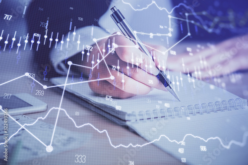 A woman hands writing information about stock market in notepad. Forex chart holograms in front. Concept of research. Multi exposure