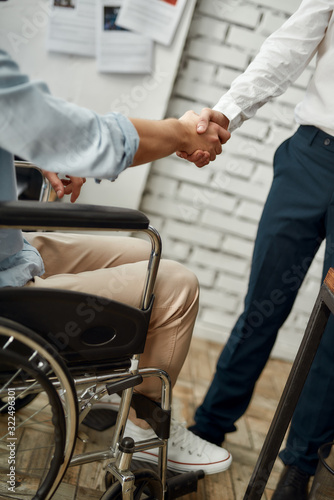 Cropped view of male office worker in a wheelchair shaking hand to his colleague while working together in the creative office. Disability concept