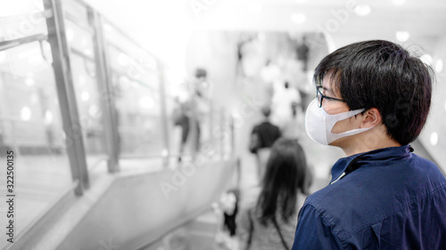 Asian man wearing surgical face mask in subway station with crowded people walking pass. Wuhan coronavirus (COVID-19) outbreak prevention in public area. Health care and medical concept