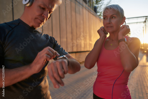 Sporty middle-aged couple in sport clothing adjusting headphones and setting their playlist while exercising together outdoors