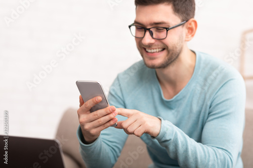 Joyful Guy Using Smartphone Texting Sitting On Couch At Home