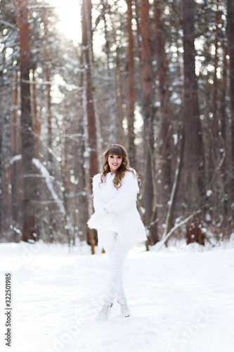 A beautiful pretty young girl with curly hair and a snow-white smile in a white warm fur coat is walking and fooling around in the winter forest against the background of snow and trees, enjoying