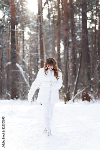 A beautiful pretty young girl with curly hair and a snow-white smile in a white warm fur coat is walking and fooling around in the winter forest against the background of snow and trees, enjoying