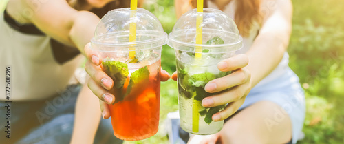 Two hipster girls are laughing and drinking summer cocktails outdoors in the green grass. Cold non-alcoholic drinks with ice to go. Mojito and strawberry lemonade. Happy lifestyle for vacations.