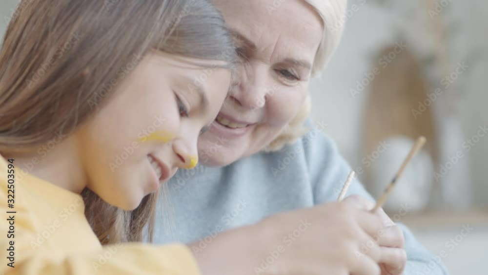 Cheerful little girl with yellow gouache on nose and cheek smiling and chatting with joyous grandmother while painting Easter egg together at table Cheerful little girl with yellow gouache on nose and cheek smiling and chatting with joyous grandmother while painting Easter egg together at table