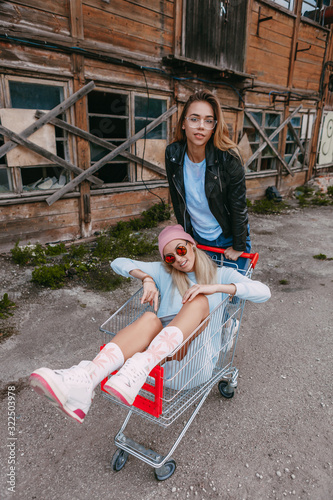 A long haired woman wearing a powder blue dress, pink woollen hat is chilling with her female friend, who is sitting on a shopping trolley, wearing a black leather jacket and blue jeans, in a derelict