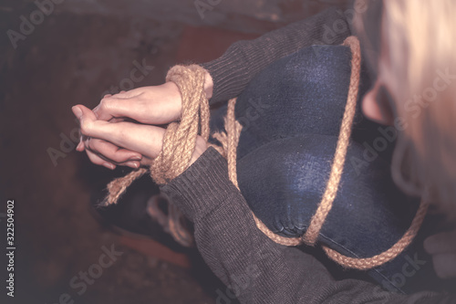 blonde girl with bound hands and feet is sitting in the basement, covering her face with her hands and crying. Concept of violence, kidnapping photo